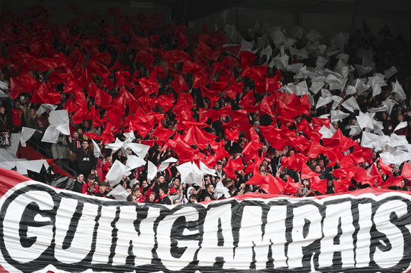 Behind a banner that reads GUINGAMPAIS there are numerous football supports holding white and red flags forming a red diagonal stripe bordered by white.