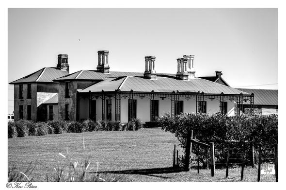 A dramatic black and white photo of Highfield House, a historic colonial building in Stanley, Tasmania.

The front of the house features a long verandah with decorative iron lacework and several tall, prominent chimneys rising above the corrugated iron roof.

A two-story wing is visible on the left. In the foreground, there is a grassy lawn, some low bushes, and a small wooden fence/gate. The sky is bright and clear.

The photo is signed by Kev Peirce.