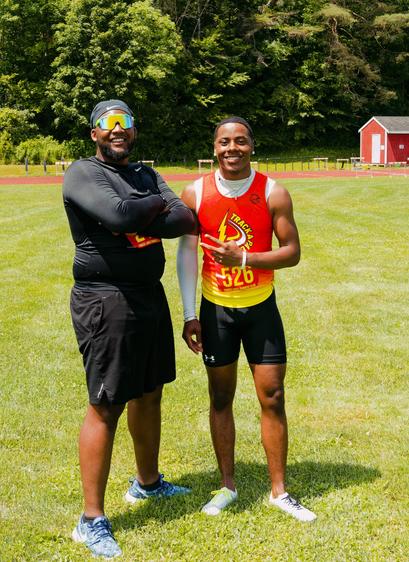 Two black men,  to the left wearing all black clothes and a black cap and dark sunglasses with his arms folded,  to the right wearing black shorts and a jersey with a yellow-to-red gradient pointing at the other with two fingers,  both smiling on a green grass field color graded to look a bit like fall all this is the summer and you can see a red track in the far distance together with a shed and a wall of trees.