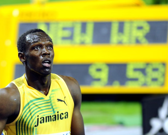 Usain Bolt is wearing a yellow shirt with green lines with the word jamaica and a puma logo and looks shocked,  behind him there is a yellow sign with the digital lets NEW WR 9.58