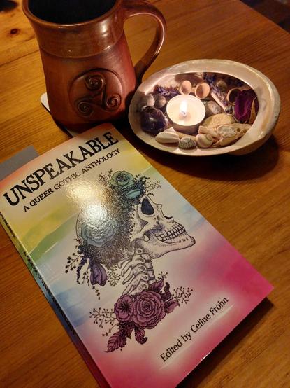 On a wooden table is a copy of 'Unspeakable: A Queer Gothic Anthology (edited by Celine Frohn). The book cover features a drawing of a skull and neck bones with flowers against a pale rainbow background. Next to the book is a large sea shell with a lit tea light and various bits of plants, shells, and bones in it. There is a brown mug with a triple spiral also with those things.