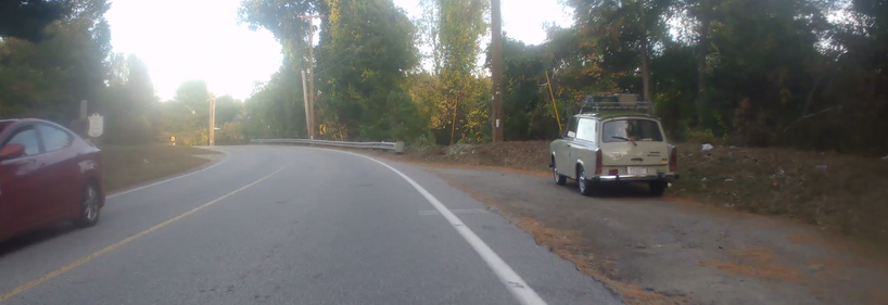 The picture is of a main road from the perspective of a handlebar camera. On the left an oncoming red car is passing, and on the right, an old Trabant is parked with DDR sticker and old DDR flag. I