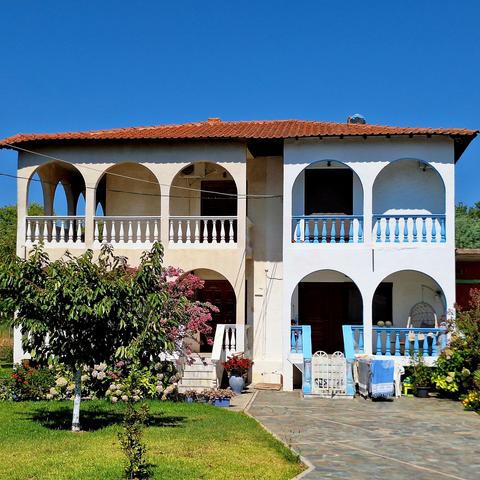 South-west facade of a countryside house made of masonry and concrete. The two storey house consists of a central block, surrounded on three sides (strangely not the South side) by arched verandas with balustrades consisting of small concrete columns. The building is slightly elevated to protect it from flooding and covered by a relatively flat hip roof. The left side is painted beige, while the right side is white and blue, showing that the small building houses two separate units.