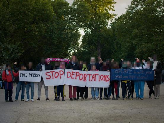 A group of people standing on Oranienplatz, some raising fists, some holding signs saying "free yerro", "stop deportations" and "oplatz is with you! You are not alone!"