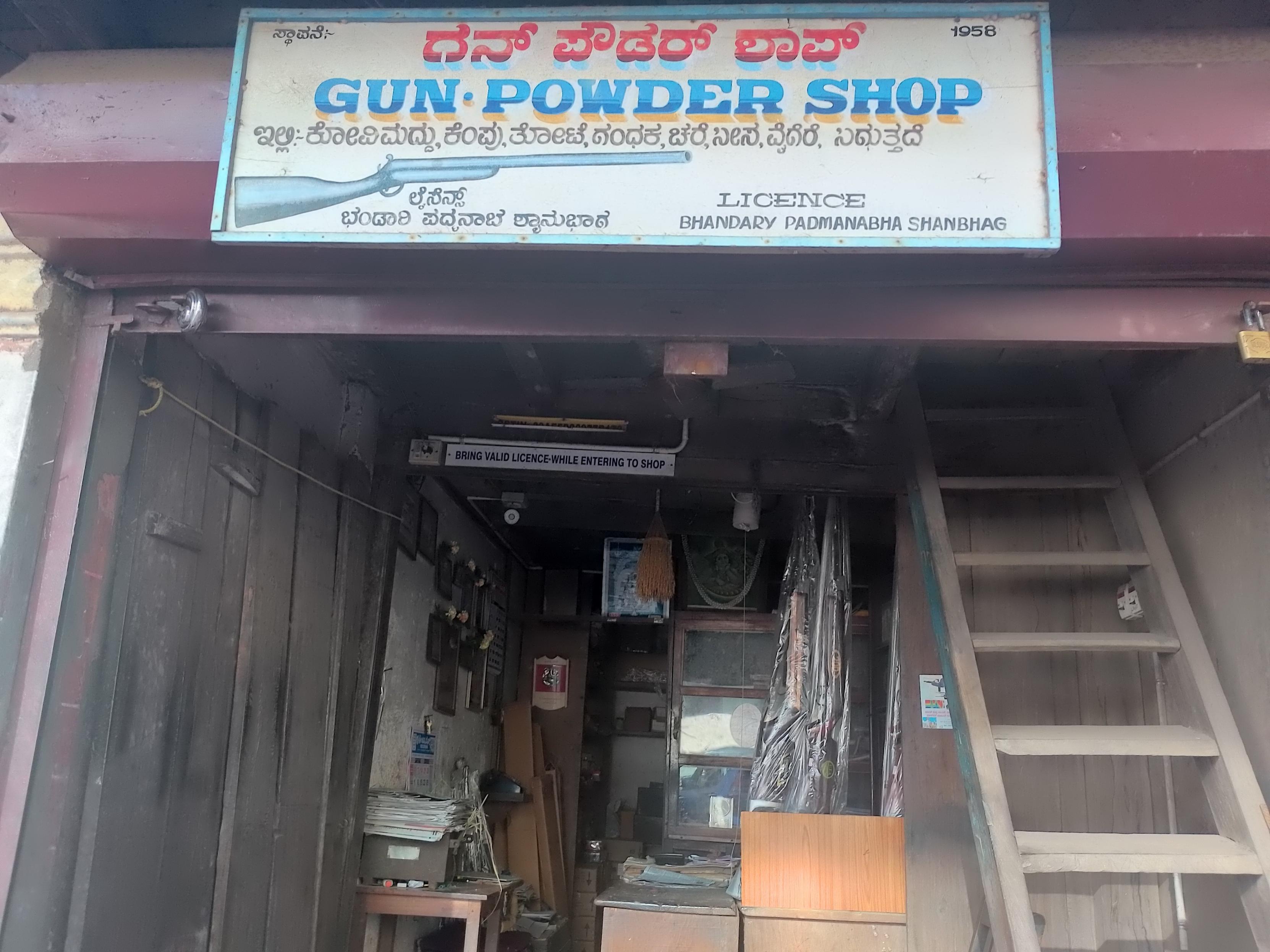 This is a photograph of the exterior of a small shop. A signboard outside the shop reads, "Gun powder shop. 1958. License Bhandary Padmanabha Shanbag." There is an illustration of a rifle and some text in Kannada on the signboard. One can see the old wooden door of the shop, a staircase and some rifles. This is a photograph of the exterior of a small shop. A signboard outside the shop reads, "Gun powder shop. 1958. License Bhandary Padmanabha Shanbag." There is an illustration of a rifle and some text in Kannada on the signboard. One can see the old wooden door of the shop, a staircase and some rifles.