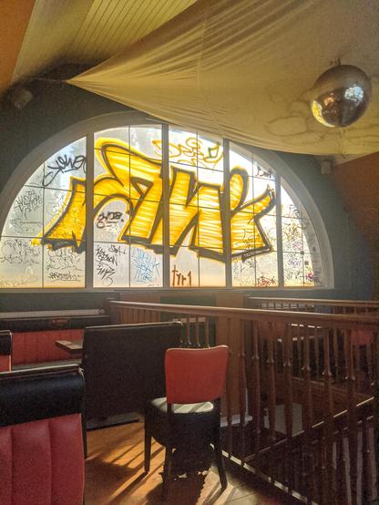 A kebab shop attic with semi-circular window. On the window a big, yellow graffiti tag and bunch of other, smaller graffiti tags. Leather chairs and sofas, wooden stairs leading down, in the upper right corner a disco ball. Everything is soaked up in warm, soft, autumn light