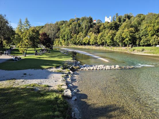 A picturesque riverside park on a bright, sunny day, featuring a gently flowing river with clear, shallow waters and a rocky shoreline. Lush green grass and trees in vibrant autumn colors create a serene and inviting atmosphere. People are seen relaxing and enjoying the beautiful surroundings, while a charming building or castle perched on a hill in the background adds a touch of historical elegance to the scene. The image captures the perfect blend of nature, relaxation, and scenic beauty.