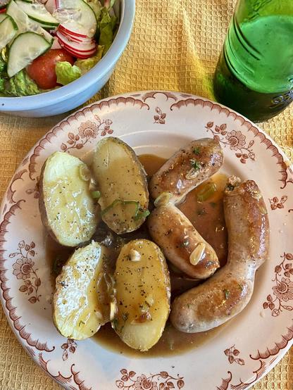 A plate contains two boiled potatoes, partially sliced open and topped with sauce, alongside several sausages. A bowl of fresh salad with lettuce, radishes, and cucumbers is in the background, along with a green bottle of beer. 