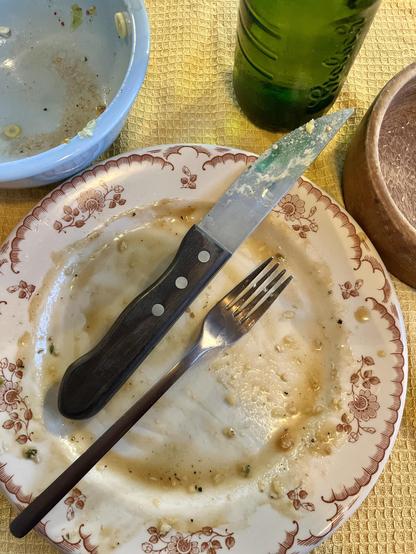 A used plate with remnants of food, a knife, and a fork. Additionally, there is an empty blue bowl and a green glass bottle visible on a textured yellow tablecloth.