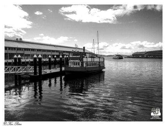 A black and white photograph of an older, two-deck ferry boat tied up to a wooden dock on the Hobart waterfront.

The boat is centered and reflects clearly in the calm water. A long, low warehouse building stretches across the background to the left.

In the distance to the right, another ferry can be seen, and rolling hills form the horizon under a partially cloudy sky.