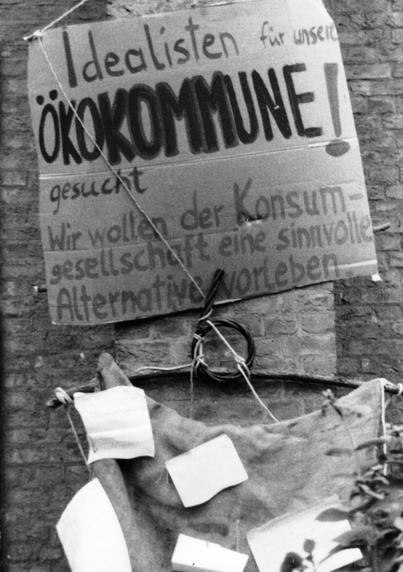 Ein handgemaltes Pappschild mit der Aufschrift: Idealisten für unsere Öko-Kommune gesucht! Wir wollen der Konsumgesellschaft eine sinnvolle Alternative vorleben.
