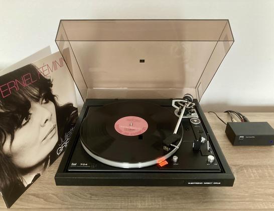 Auf dem Foto sihet man einen Dual CS-704, welcher das Album 'L'éternel Féminin' von Juliette Greco abspielt. Links neben dem Plattenspieler steht das Cover des Albums, auf dem Grecos Kopf zu sehen ist. Rechts neben dem Plattenspieler steht ein NAD Vorverstärker.