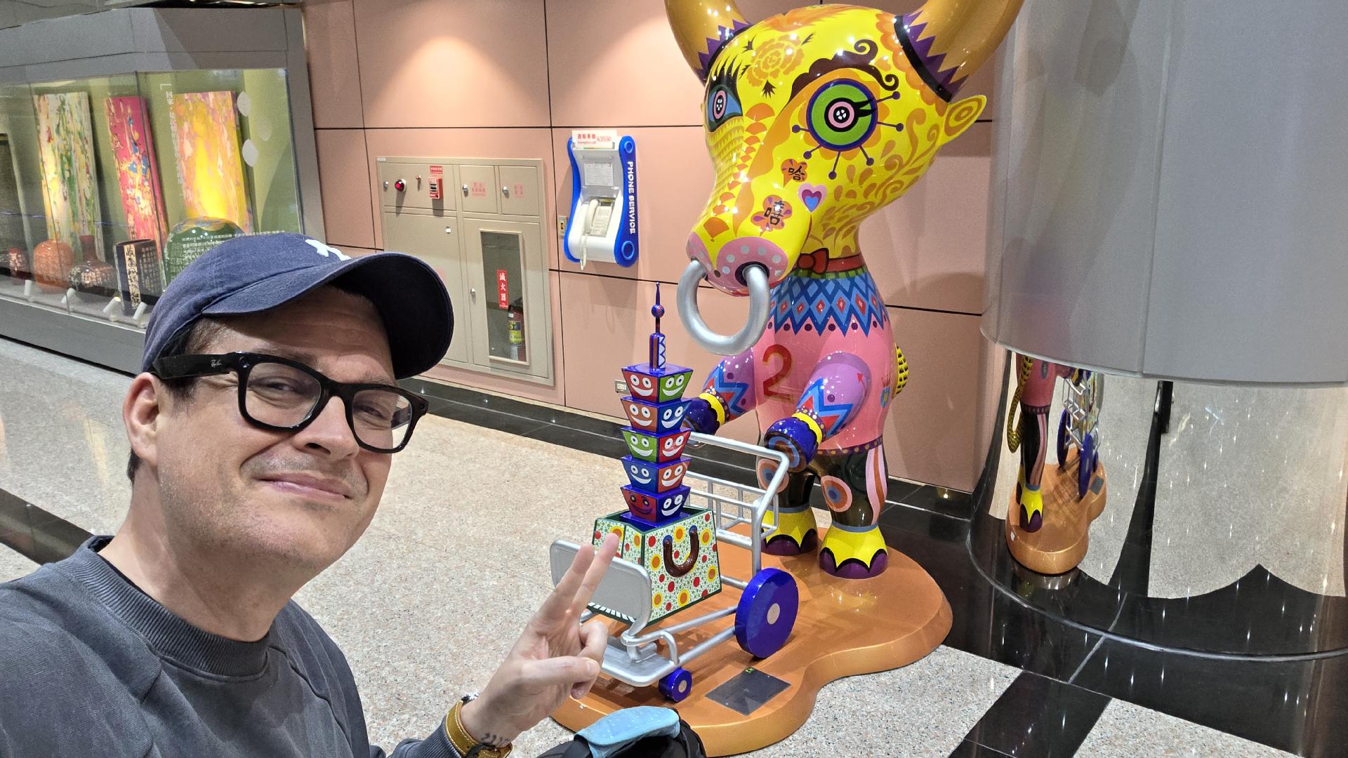 Das Foto zeigt eine Person, die ein Selfie in einer öffentlichen Halle oder einem Transitbereich (vermutlich ein Flughafen oder eine U-Bahn-Station) macht.
Im Vordergrund ist die Person mit Brille, Basecap und Sweatshirt zu sehen, die lächelnd in die Kamera schaut und mit der rechten Hand ein Peace-Zeichen macht.
Im Hintergrund steht eine bunte, kunstvoll bemalte Skulptur in Form eines Tieres – es erinnert an ein stilisiertes Nashorn oder Elefant mit großen Ohren, Ring durch den Rüssel und kräftigen, geometrischen Mustern in knalligen Farben (gelb, pink, blau, grün, orange). Die Skulptur zieht einen kleinen Wagen mit mehreren übereinandergestapelten, bunten Schalen mit Gesichtern darauf.
An der Wand daneben hängen ein Münztelefon und mehrere Sicherungskästen, während im Hintergrund Vitrinen mit farbenfrohen Kunstwerken und Keramiken zu sehen sind.
👉 Die Szene wirkt freundlich und verspielt – ein Moment zwischen Alltagslogistik und öffentlicher Kunstinstallation, eingefangen mit einem Augenzwinkern.