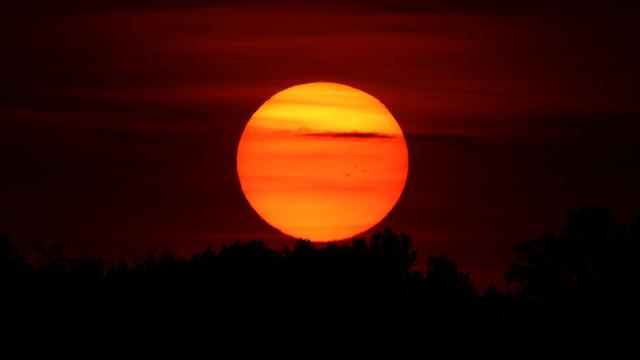 Photograph of a large Sun at sunset with yellow and orange stripes from clouds and several small sunspots visible. The sun is setting into a treeline with a dark red sky behind.