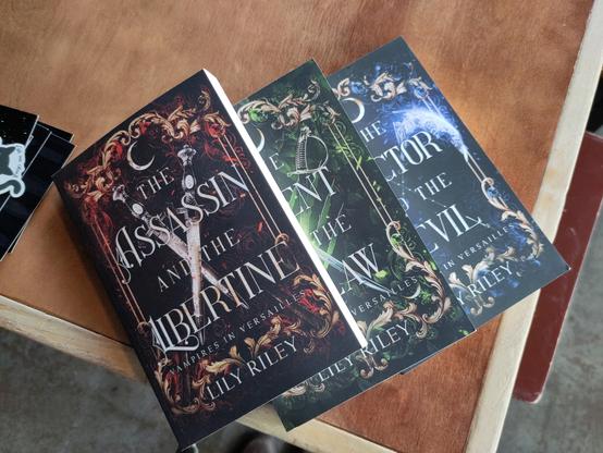 Photo of three novels on a restaurant table: The Assassin and the Libertine, The Agent and the Outlaw, The Doctor in the Devil. This is a trilogy (or maybe the first three of a series?) by Lily Riley. Vampires in Versailles sound fun to me. 