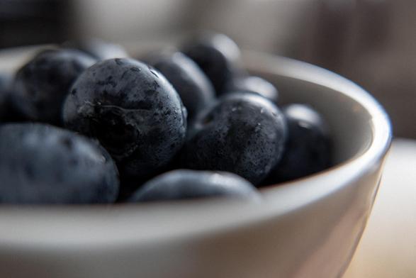 A close up colour photograph of some large blueberries in a white ceramic bowl.  Some small water droplets are visible.  The scene is lit from right side, slight reflections are bouncing off of the bowl and the berries.