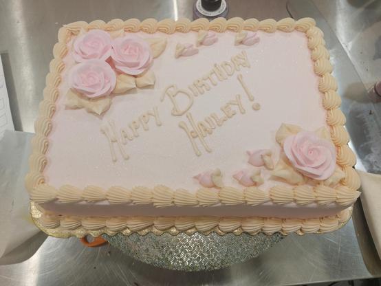 A small sheet cake covered in pastel pink icing, with pastel orange shell borders. There are three pink roses in the upper left corner, and one in the lower right, they all have several orange leaves around them; three rosebuds trail off from the roses in either corner. "Happy Birthday Hailey" is printed diagonally across the center of the cake in the pastel orange color as the borders.
