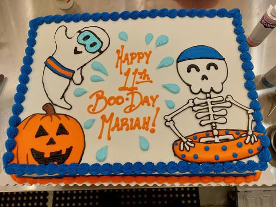 A small sheet cake with white icing and royal blue and orange borders. The lower left corner features a Jack-o-Lantern, above it is a ghost wearing a swim mask and a blue and orange waistband. Across from them is a skeleton with a blue swim cap and orange inner tube with blue polka dots. "Happy 11th Boo Day Mariah" is printed in orange at the center and surrounded by water droplets. The party theme was a Halloween pool party.