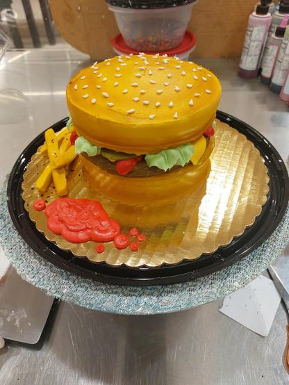 A small two-layer round cake decorated to resemble a hamburger, complete with fondant French fries and a blob of red icing ketchup. The burger itself is chocolate frosting, the toppings are visible and include frosting as lettuce, ketchup, and mustard, as well as a thin slices of fondant as pickles and cheese. Numerous sesame seeds (also made from frosting dots) are scattered across the top of the bun.
