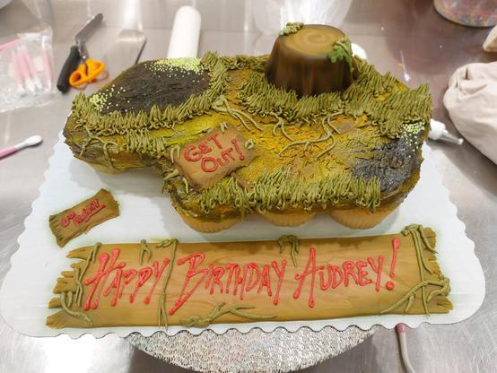 A cupcake pull-apart cake airbrushed to resemble a grassy swamp (the party had a Shrek theme). There are three wooden fondant signs reading "Get out," "Go away" and "Happy Birthday Audrey!" A tree stump, made from an upside-down cupcake covered in fondant, rises out of the olive-green grass and vines.