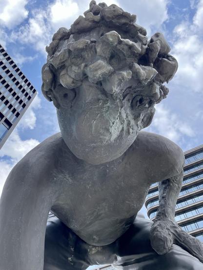 A large, abstract bronze sculpture depicting a figure with curly hair crouching down, set against a backdrop of modern buildings and a partly cloudy sky.