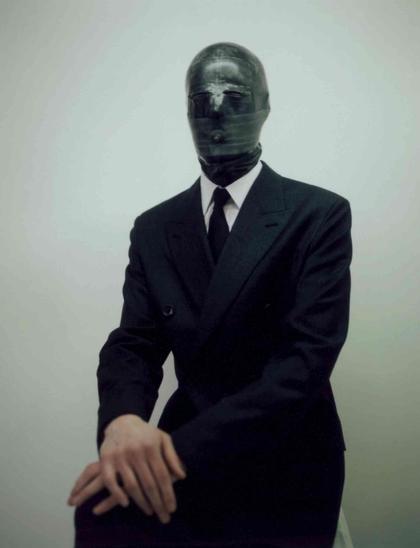 Figure in a dark suit and white shirt seated against a pale background, with their face completely covered by a dark, possibly rubber or latex, mask featuring a small breathing hole.