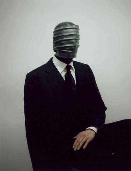 Man in a dark pinstripe suit and tie wearing a dark, horizontally ridged, featureless mask against a pale gray background.