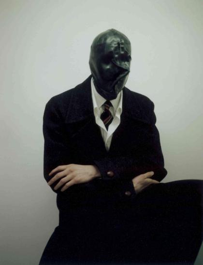 Figure wearing a dark, tight-fitting facial covering, a white shirt, striped tie, and black overcoat sits against a plain light background.