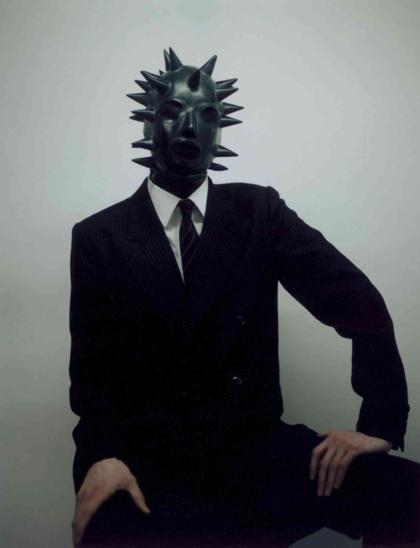 Figure in a dark, spiked rubber mask and a black pinstripe suit sitting against a plain light gray background.