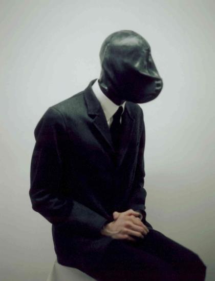 A person in a dark suit and white shirt sits with clasped hands, wearing a featureless black latex or rubber mask against a pale gray background.