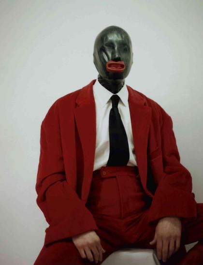 Person wearing a black latex hood with red lips and an oversized red suit over a white shirt and black tie against a light background.