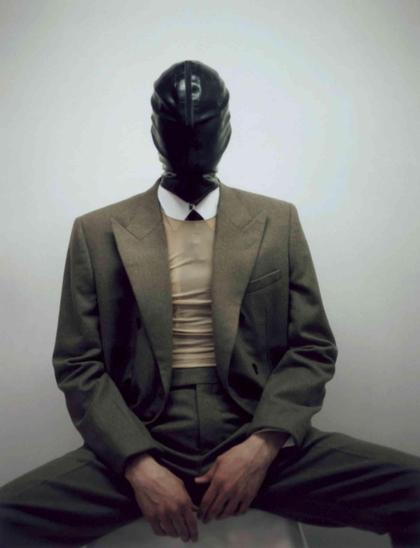 Figure in a tailored olive green suit and beige ribbed top wears a shiny black leather hood against a pale background.