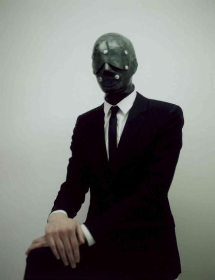 Person in a black suit and tie wearing a dark, studded, leather-like mask against a light gray background.
