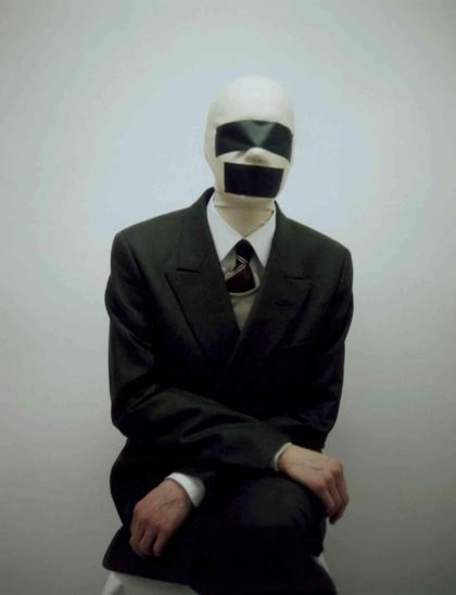 Person in a dark suit and white head covering with black tape obscuring his eyes and mouth sits against a plain light gray background.