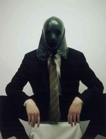 Person wearing a dark suit and striped tie sids with their legs wide open while obscuring their face with a dark, smooth hood or mask against a plain white background.