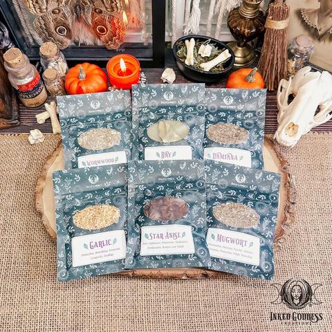 Six small pouches of dried herbs from the Samhain Herb Set are arranged on a raw wood slice. The pouches are about the size of a seed packet and have a clear window on the front. The label on each pouch features the name of each herb and its potential magickal uses: Wormwood, Bay, Damiana, Garlic, Star Anise, and Mugwort. This is an exclusive product from Inked Goddess Creations.