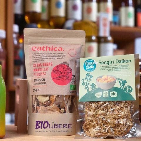 Una vista cercana muestra dos paquetes de alimentos orgánicos deshidratados colocados sobre una superficie de madera clara, con un fondo borroso de estantes de tienda llenos de botellas y tarros.

A la izquierda, un paquete vertical de papel kraft de la marca "eathica." Contiene "SETAS ROSAS, AMARILLAS Y GRISES ECOLÓGICAS DESHIDRATADAS" de 25g. El envase tiene una ilustración circular de setas rosadas y logos que indican "PRODUCTO LOCAL" y certificación orgánica de la UE. Una ventana transparente en la parte inferior del paquete permite ver las setas deshidratadas de color beige y marrón.

A la derecha, un paquete de plástico transparente de la marca "Terra Sana" para "Sengiri Daikon" (rábano blanco seco deshidratado). La parte superior del paquete es una etiqueta verde y blanca que muestra un paisaje asiático con árboles, una pagoda y figuras, junto con una imagen de un bol de daikon preparado. Incluye logos de "VEGAN", "SOURCE OF FIBRE" y "BIO", además de un sello japonés de "Authentic Taste". El rábano blanco deshidratado, de aspecto fibroso y claro, es visible a través del plástico transparente.