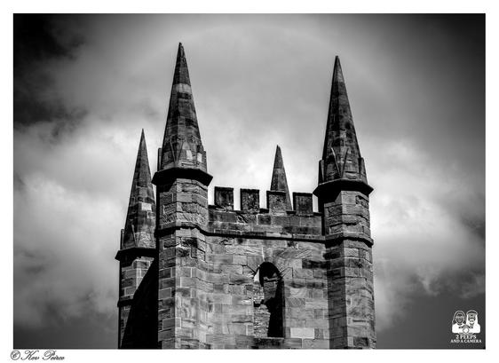 A dramatic black and white photograph of the top of the ruined Anglican Church at Port Arthur, Tasmania.

The stone tower, with its distinctive spire-like pinnacles and crenellated top, is silhouetted against a cloudy, bright sky, creating a high-contrast and moody effect.

A halo effect is visible around the main structure against the clouds. The photograph is signed by Kev Peirce.