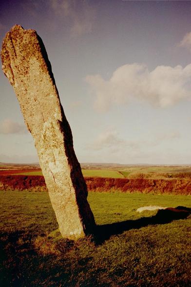 The NE Piper, a prehistoric standing stone in West Penwith, Cornwall. Standing in a green field and casting a very long winter's shadow. In the middle distance a rusty red hedge crosses below a series of gentle rolling hills. The sky above is pale blue with several clouds.