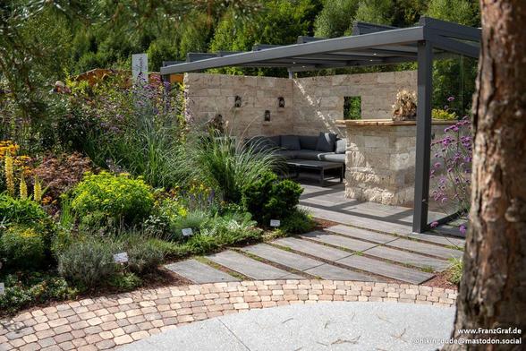 A serene outdoor patio is featured in this image, showcasing a beautiful stone wall and a stone structure. The overall color scheme includes dominant shades of grey and white, with an accent color of 95A922. A lush tree and various plants can be seen throughout the landscaping, adding a touch of nature to the scene. A bench provides a cozy spot to sit and enjoy the surroundings. The patio exudes tranquility and charm, making it a perfect spot for relaxation and unwinding. The image also include…