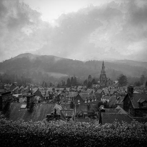 A black and white landscape featuring a village with a mix of houses and a prominent church steeple. The scene is set against a backdrop of misty hills and overcast skies, conveying a tranquil, moody atmosphere.