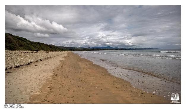 A wide, horizontal photo of a sandy beach stretching into the distance under a dramatic, cloudy grey sky.

To the left, the beach meets a bank covered in rough stones and scrubby green vegetation.

The sand is wet and reflective where it meets the incoming gentle waves on the right. In the distance, a coastline and rolling hills or mountains are visible under the clouds.

The photo is signed 'Kev Peirce'.
