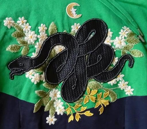 Close up of a green and navy blue hoodie front, with a black snake patch, green and gold foliage and white flowers. The snake is outlined with dark grey seed beads.