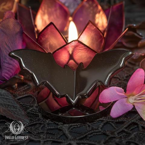 An example of the "Obsidian Flying Bat" product stands on a dark stone disc. The glass is carved into the shape of a flying bat, wings spread and bent in a V shape. Presented by Inked Goddess Creations.