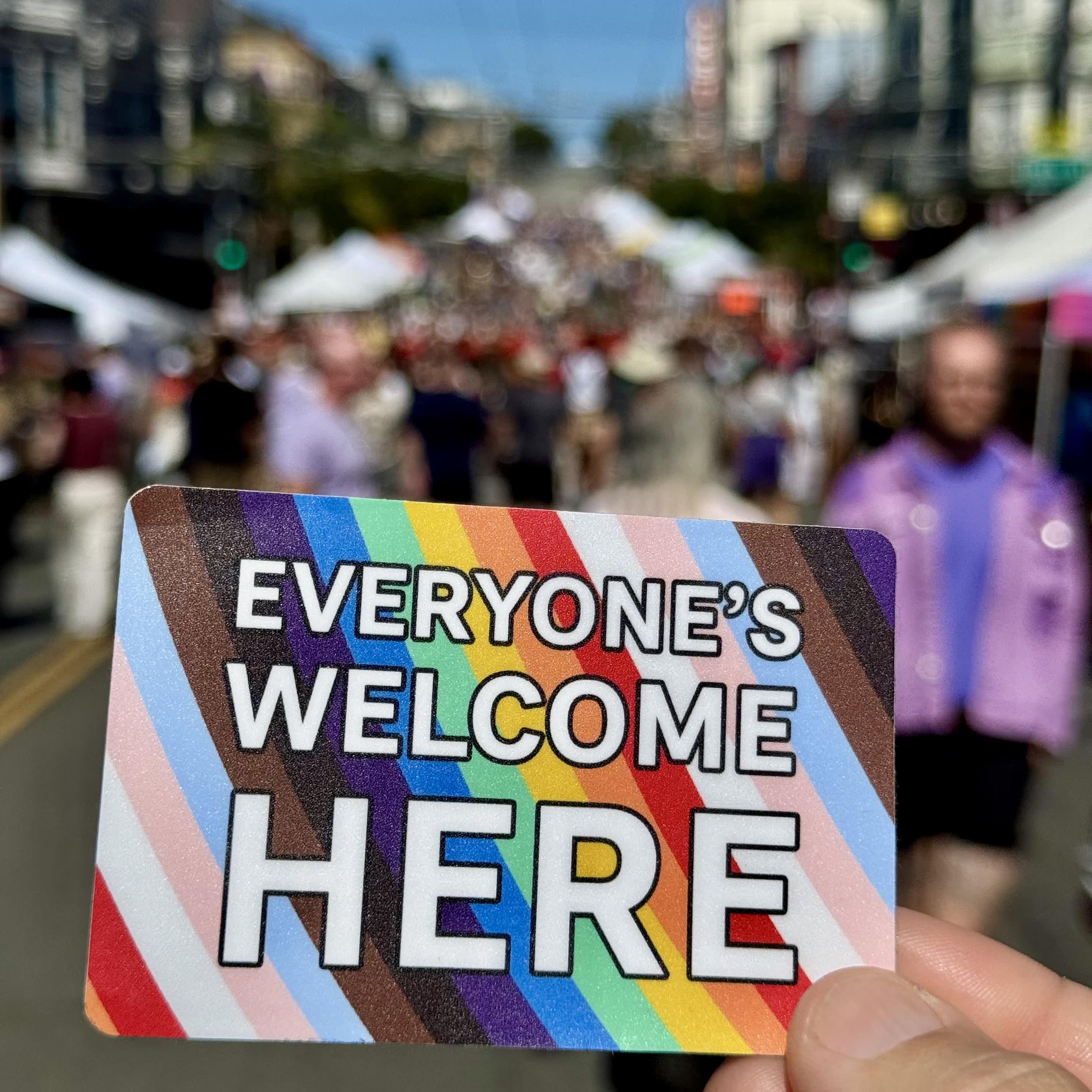 A sticker in the foreground with a large art fair on Castro Street in the background. The sticker says "Everyone's Welcome Here" in white letters in front of a LGBQTIA+ Pride Flag.