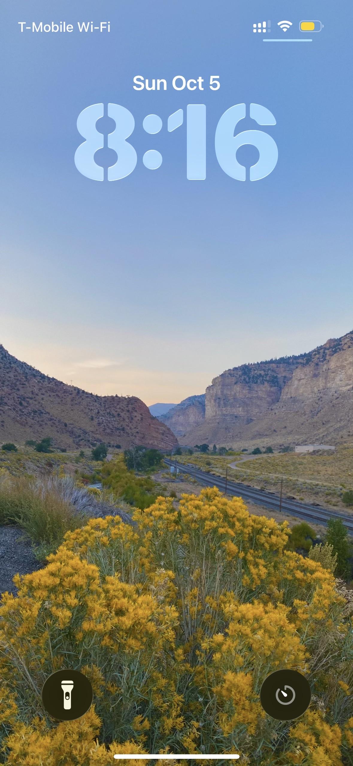A scenic view of a landscape featuring yellow wildflowers in the foreground and rocky hills in the background. The sky is clear with a soft blue gradient, and the time displayed is 8:16 on Sunday, October 5.