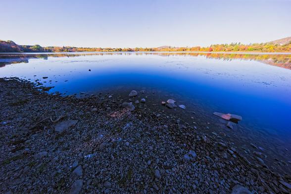 At the very bottom a beach with irregular bits of gravel,  above that a body of water that is dark blue at the bottom and white with reflections near the horizon and has a landscape that curves up away from the horizon away from the center with sky that doesn't quite as dramatic as its reflection