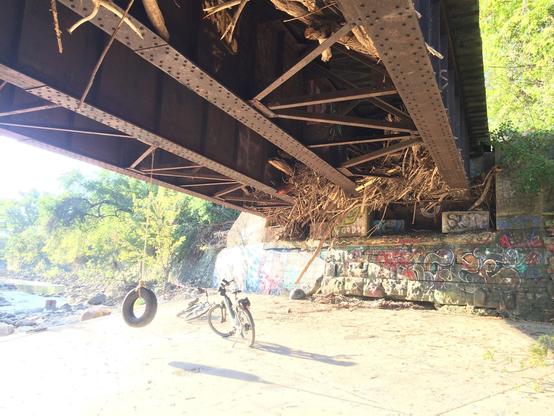 Two bikes and a tire swing under a rail bridge that has branches and debris stuck under it.