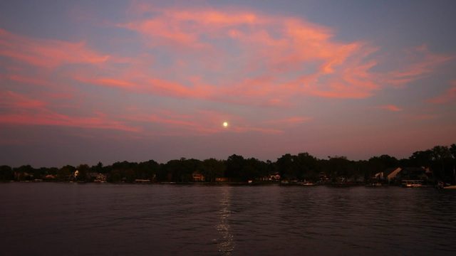 Photo of a nearly full moon rising over a lake surrounded by pink clouds.