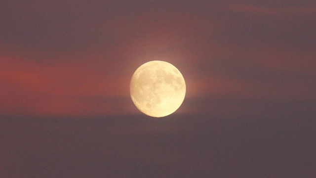 Photo of a medium-large nearly full moon surrounded by pink clouds.
